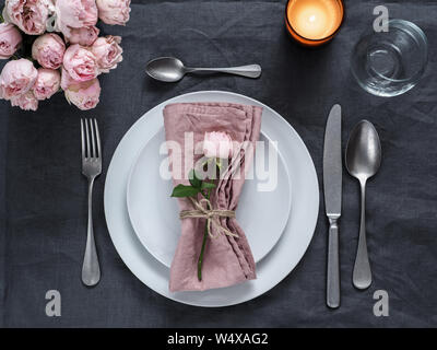 Beautiful table setting with candle on gray linen tablecloth. Festive table setting for wedding dinner with pink spray rose and pink napkin on plate. Holiday dinner with white plates Stock Photo
