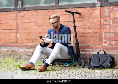 A Handsome Businessman Sitting On Electric Scooter Using Cellphone Near Brick wall Stock Photo