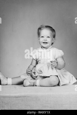Full length, seated portrait of a smiling  young girl wearing a long dress and laced shoes, sitting on a fabric surface, legs facing to the side and both hands held up, 1960. () Stock Photo