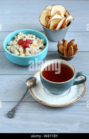 Breakfast: oatmeal with fruit pieces and candied strawberries, pear and apple chips in small bowls and a cup of black tea on a gray wooden table. Clos Stock Photo