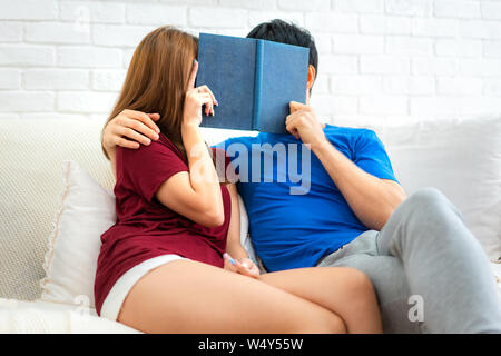 Spending nice time at home. Beautiful young loving couple reading book and smiling at couch in living room at home. Stock Photo