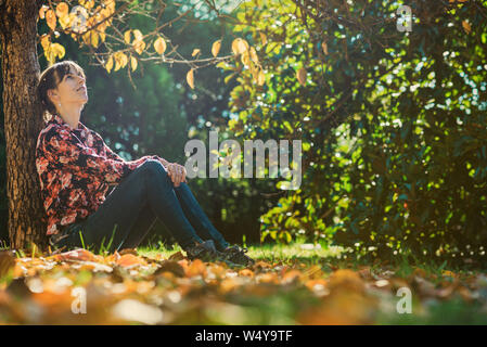 Young woman sitting under an autumn tree leaning on the tree trunk enjoying and relaxing. Stock Photo