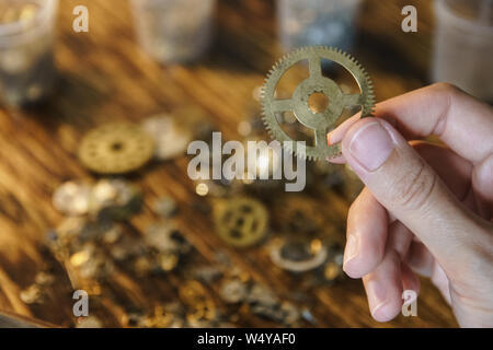 Watch repairer holds large gear. Big metal gearwheel on blurred workspace. Cogwheel on blurred background of workplace. Pinion in female hand. Old cog Stock Photo