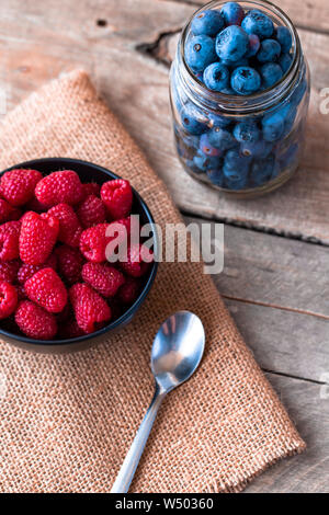 Blueberries in a jar and raspberries in a wood bowl on top of burlap with a wooden table top Stock Photo