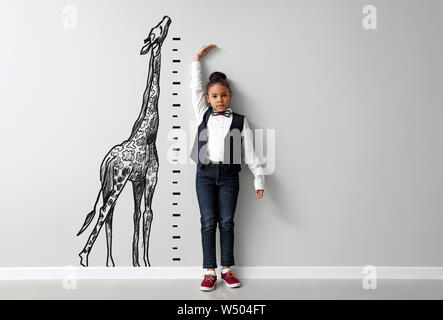 Cute little African-American girl measuring height near light wall with drawn giraffe Stock Photo