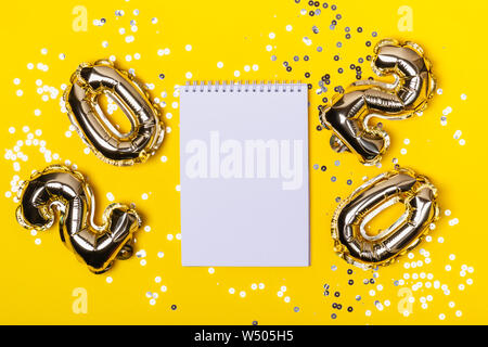 Creative layout made of gold foil balloons in shape of numbers 2020, blank notepad and confetti sparkles. Stock Photo