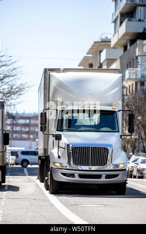 Day cab big rig white professional semi truck transporting commercial cargo in refrigerated semi trailer running on the urban city street with multile Stock Photo