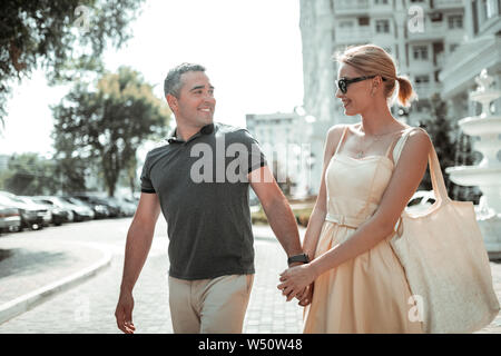Lucky man. Happy smiling man looking at his beautiful cheerful wife and holding her hand walking through the town. Stock Photo