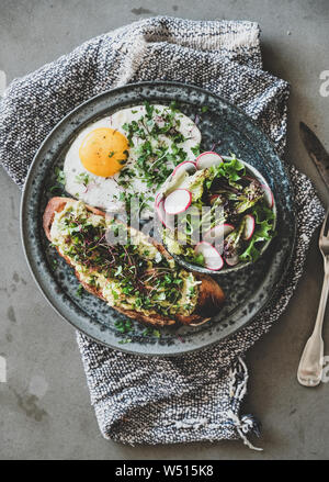 Healthy trendy breakfast plate. Flat-lay of avocado toast on sourdough bread with herbs and sprouts, fried egg with coriander and salad over grey conc Stock Photo