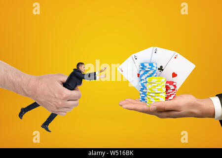 Man's hand holding tiny businessman who is reaching out with both hands for cards and chips on another man's palm on amber background. Stock Photo