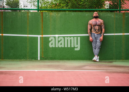 Attractive man with tattoos and beard wearing training pants standing on tennis court looking front Stock Photo