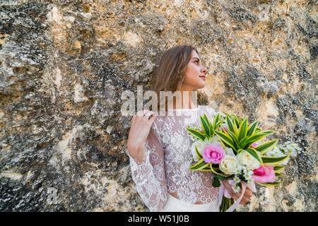 Beautiful bride in white dress holding flowers in one hand and touching hair with other, leaning against cliff wall in Bali Stock Photo