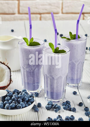 close-up of blueberry frothy milkshake of coconut milk in three glasses with straws. cracked coconut and jug with milk on a white wooden table, horizo Stock Photo