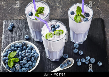 close-up of blueberry frothy milkshake of coconut milk in three glasses with straws on a black slate tray on a concrete table, horizontal view from ab Stock Photo
