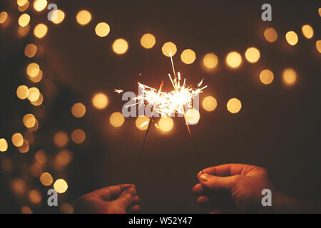 Happy New Year. Glowing sparklers in hands on background of golden christmas tree lights, couple celebrating in dark festive room. Space for text. Fir Stock Photo