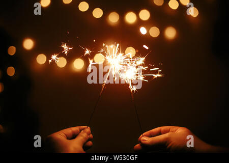 Happy New Year. Glowing sparklers in couple hands on background of golden christmas tree lights, family celebrating in dark festive room. Space for te Stock Photo