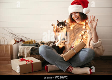 Stylish happy girl in santa hat  and cute cat waving hand and paw in festive christmas lights on background of modern presents. Young hipster woman pl Stock Photo