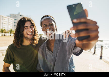 Two young men standing together outdoors taking a selfie and smiling. Best friends taking a selfie while standing on a seaside road on a summer day. Stock Photo