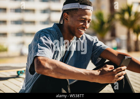 Smiling african american man sitting outdoors and reading text messages on his smart phone. Young skateboarder smiling while using mobile phone. Stock Photo