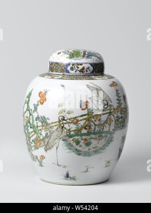 Ovoid covered jar with two cranes in a fenced garden, Egg-shaped porcelain covered pot, painted on the glaze in blue, red, green, yellow, eggplant and black. On the wall a fenced garden with two cranes, one standing and one on the fence, by a pond with lotus plants, a rock and a flowering tree (magnolia). Plants that bloom around the pond (chrysanthemum, beautiful carnation, peony, aster, lily) and two flower pots with pine, bamboo and orange. A parrot on a rack in the magnolia tree. On the right two flying birds. Napkin work on the shoulder interspersed with flower sprays in cartouches. Stock Photo