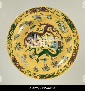 Dish with pearl chasing dragons on a yellow ground, Porcelain dish with deep wall and flat border, painted in underglaze blue and on the biscuit green, yellow, eggplant and black. On the shelf in an engraved medallion, two pearl chasing dragons between flames and clouds, eight flower branches on the wall (including lotus, peony, prunus, chrysanthemum), the border with dragons, the rear of the edge with five flying cranes among clouds, the back of the wall with dragons. Marked on the underside with the six-character mark of emperor Kangxi in a double circle. Email sur biscuit., anonymous, China Stock Photo