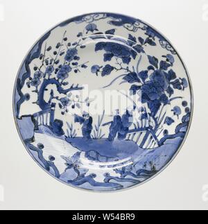 Dish with three figures in a water landscape with flowering plants, Porcelain dish, painted in underglaze blue. On the front a continuous representation of three people in a landscape on the banks of a river. One man has a role in his hand, another is close to the water and points to the other side. To the right of the group a large peony plant behind a gate, on the left a prunus tree behind a fence. Five proen on the bottom. Old label on the bottom with 'P [?] 8031 / Imari / - 1700 / f 475'. Arita, blue and white., anonymous, Japan, c. 1675 - c. 1725, Edo-period (1600-1868), porcelain Stock Photo