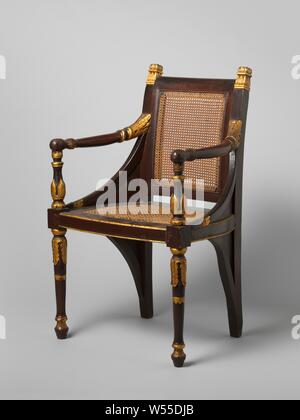 Armchair, Empire model, with back and seat made of cane, painted red and gilded and heightened with gold, Two armchairs (A and B) of a tropical wood (djati?), Painted red, parts gilded and heightened with gold, the back and the loose seat frame covered with reeds. Empire model with straight back, slightly arched back, widening seat with curved sides, baluster-shaped front legs and arm struts, curved, spherical-ended arms and dense cheeks with sunken fields, which are repeated below the seat in mirror image and in smaller format. Sculpted with acanthus and palmet gold motifs. Mutual differences Stock Photo