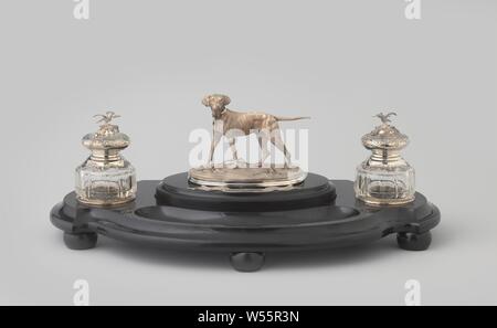 Set on scalloped frame with dog, The scalloped black marble frame, with a straight back, has an S-shaped curved wall and rests on five flattened ball legs. In the top, a round opening has been saved on each short side, covered with a silver ring with a profiled, slightly raised edge. The two cylindrical jars of colorless, cut glass fit, the rejuvenating neck of which has a silver frame. With the inkwell it has a round opening at the top, with a sand spreader a star made up of round holes. The lower edge of both frames is decorated with C-volutes, rocailles and flowers. The silver, flattened Stock Photo