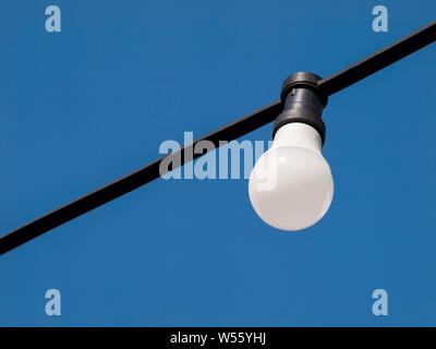 A one single white simple lightbulb on blue sky background hanging from a black power cord closeup, light turned off. Energy saving, illumination Stock Photo