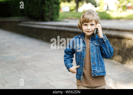 Cute blond guy, school year fun running time on the street and holds a smartphone in his hands. Open portrait of a boy in a city Park . The child is w Stock Photo