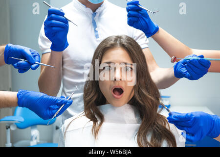 surprised young brunette woman patient sitting in dentist chair surrounded by hands in blue gloves with medical instruments Stock Photo