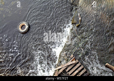 A tire and a wooden pallet have been discarded on a rocky surface in shallow rushing water at the edge of a river, and are viewed from above. Stock Photo