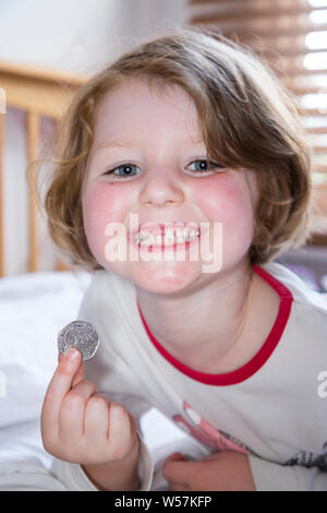 Young girl smiling to reveal a missing front tooth / teeth tooth, whilst holding a 50p / fifty  pence coin in which arrived from the tooth fairy under her pillow. UK (111) Stock Photo