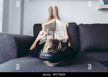 Blond boy barefoot reads book and listening music lying down. Student reading the lesson comfortably relaxed, diligent child reads a story young man p