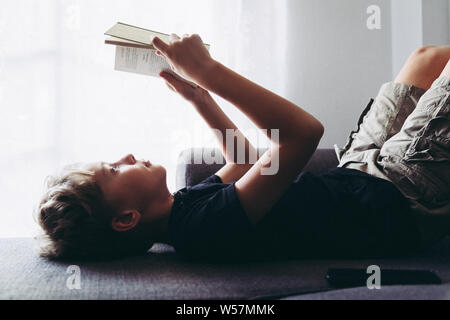 Profile view of a blond boy reads book and listening music lying down. Student reading the lesson comfortably relaxed, diligent child reads a story yo