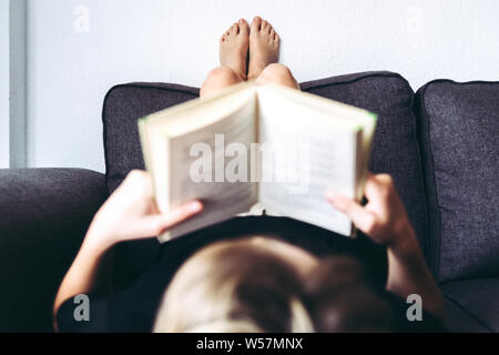 Close up view of blonde boy barefoot reading book, student lying down learn the lesson comfortably relaxed, diligent child reads a story, young man pl Stock Photo
