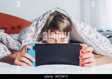 Young boy playing video games with console, impersonating himself in the game having fun with his new toy. Child using his console leaning in his bed