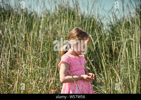 Thoughtful young girl in pink dress standing among tall grass in lush green field on Grotta island and learning wild nature Stock Photo