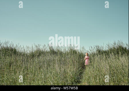 Summer holidays concept with lovely sunlit little girl in pink dress standing on trail among tall grass in lush green field on Grotta island Stock Photo
