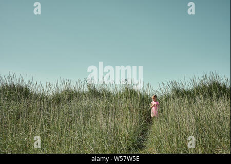 Field trip concept with pretty little girl in pink dress standing on trail among tall grass on Grotta island in summer Stock Photo