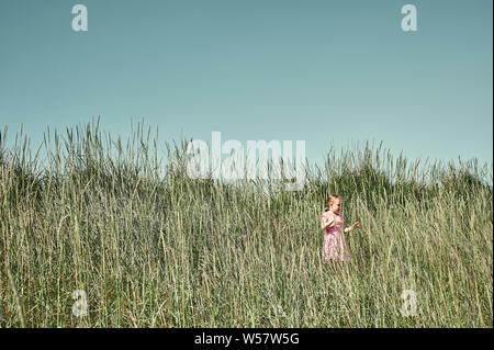Pretty young girl in pink dress walking in tall grass in lush sunlit green field on Grotta island in summer Stock Photo