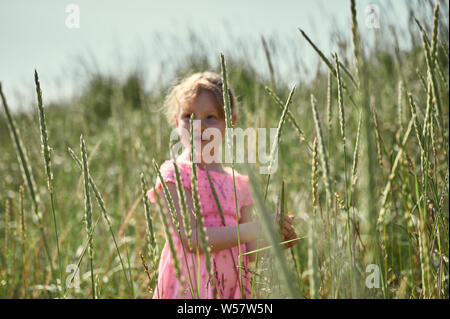 Sweet young girl in pink dress standing among tall grass in lush green field on Grotta island in summer Stock Photo