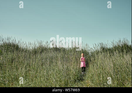 Pretty young girl in pink dress standing on trail among tall grass in lush green field on Grotta island in summer Stock Photo