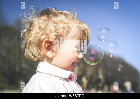 Beautiful little haired hair girl, has happy fun smiling face, pretty eyes, short hair, playing and catching soap bubbles in summer nature, dressed