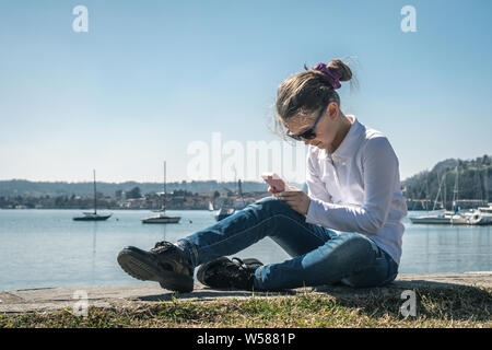 Beautiful little girl spends the festive afternoon at the playground, sitting on the shore of the lake playing with smartphone. Free time outdoors, in Stock Photo