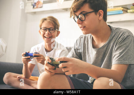 Young brothers enjoy video game at home. Smiling kids playing with console sitting on a sofa Teenager and brother playing with wireless remote control Stock Photo