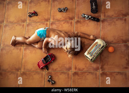 Cute little boy lying down on the floor and playing with models of cars and motorcycles at home, leisure time concept Stock Photo