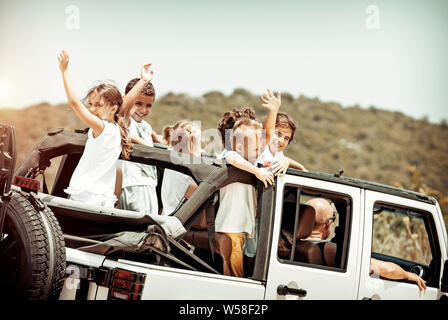 Happy kids enjoying road trip in the open-top 4x4, cheerful boys and girls having fun in the car riding along mountains, happy active summer holidays Stock Photo