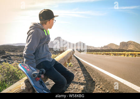 Portrait of thoughtful trendy boy with eyeglasses sitting with skateboard along a mountain road dreamily. Young man with skate looks absorbed at the s Stock Photo