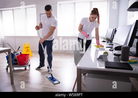 Two Smiling Young Janitor Cleaning The Desk And Mopping Floor In The Office Stock Photo